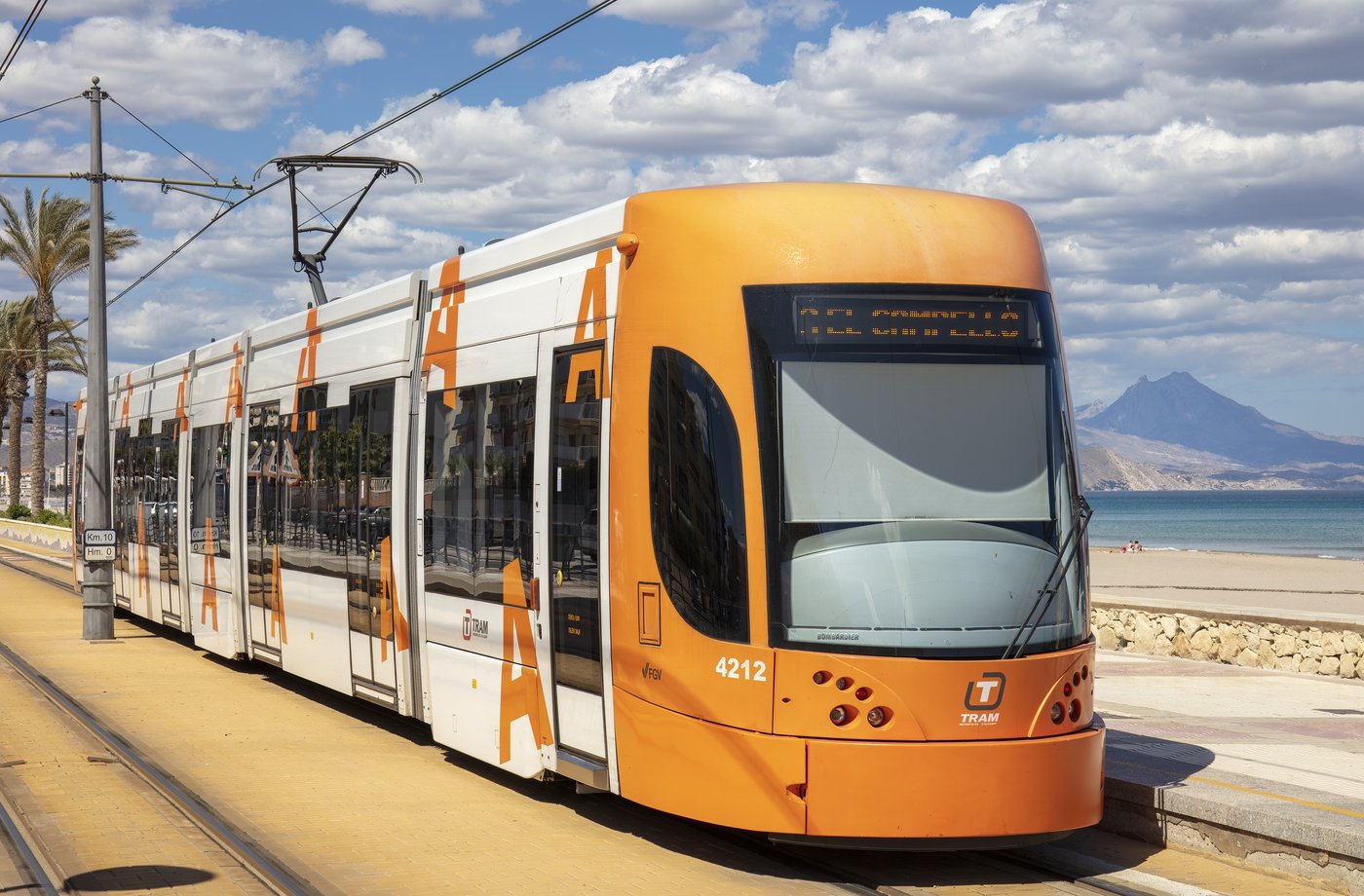 The orange Alicante TRAM running alongside the beach with the coast behind it